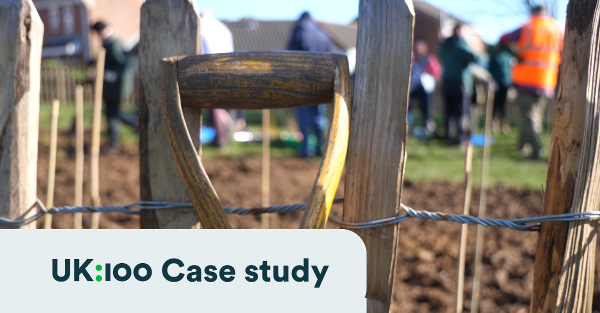 A spade lent against a wood and wire fence with a community of people out of focus in the background having just planted trees.